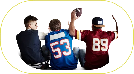 Three men dressed in sports jerseys are sitting on a sofa watching a football match on television. They are cheering enthusiastically; one of them is holding a football, while another has a foam finger.